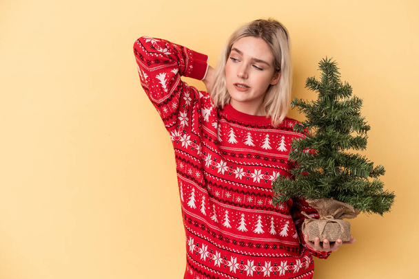 Mulher caucasiana jovem segurando uma pequena árvore de Natal isolada no fundo amarelo tocando parte de trás da cabeça, pensando e fazendo uma escolha. - Foto, Imagem