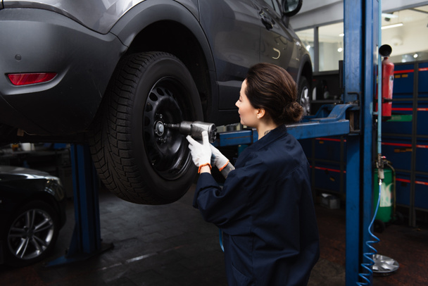 Side view of forewoman working with impact wrench on wheel of car in service  - Foto, Imagen