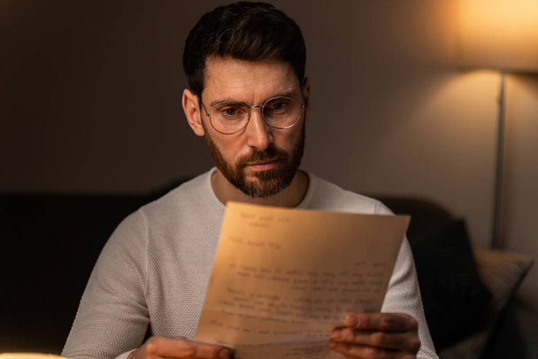 Bearded man wearing glasses reading interesting letter with serious expression while spending evening alone at home  - Photo, Image