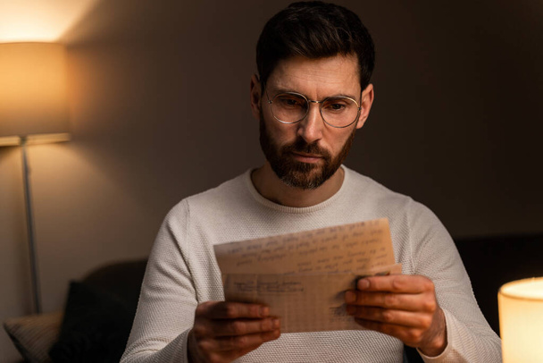 Caucasian bearded man wearing glasses sitting at the lonely room during the evening and reading interesting letter from family  - Фото, зображення