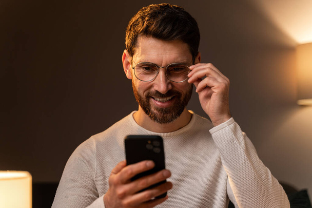 Carefree guy in home wear is sitting on cozy sofa. He is spending time with mobile phone in his home. Stock photo  - Valokuva, kuva