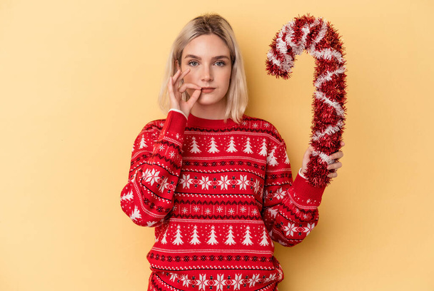 Молода біла жінка тримає велику різдвяну паличку ізольовано на жовтому тлі з пальцями на губах тримаючи секрет
. - Фото, зображення