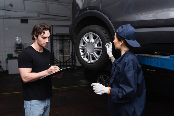 Mechanic pointing at car wheel near colleague writing on clipboard in service  - 写真・画像