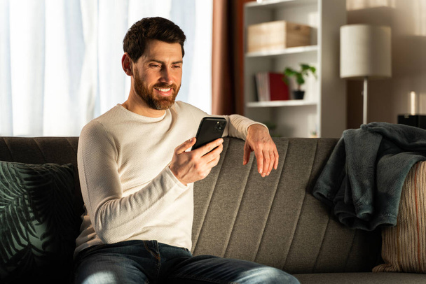 Carefree guy in home wear is sitting on cozy sofa. He is spending time with mobile phone in his flat. Stock photo  - Fotografie, Obrázek