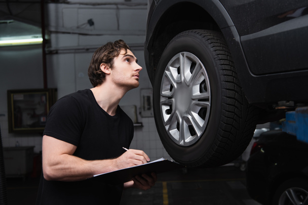 Mechanic writing on clipboard and looking at car in service  - Photo, image