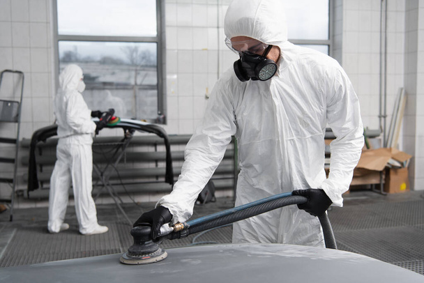 Workman in hazmat suit working with car polisher in garage  - Fotó, kép