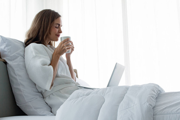 白いパジャマを着た若い白人女性がコーヒーとドリンクを飲み、自宅の寝室のベッドの上のラップトップから情報を見つけます。コーヒーを飲みながらノートパソコンで働く女性. - 写真・画像