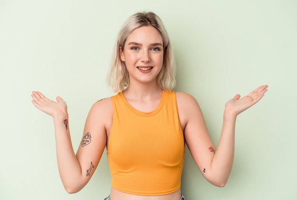 Young caucasian woman isolated on green background makes scale with arms, feels happy and confident. - Fotoğraf, Görsel