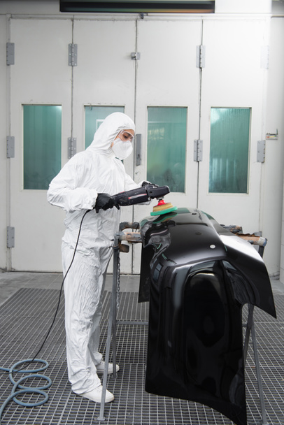 Workwoman in protective suit polishing car part in garage  - 写真・画像
