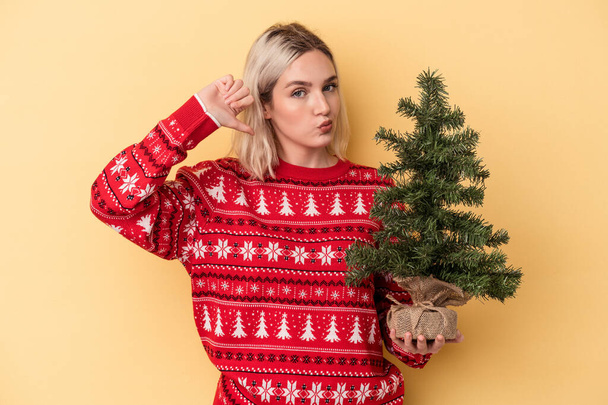 Mladá běloška drží malý vánoční stromek izolovaný na žlutém pozadí cítí hrdost a sebevědomí, příklad následovat. - Fotografie, Obrázek