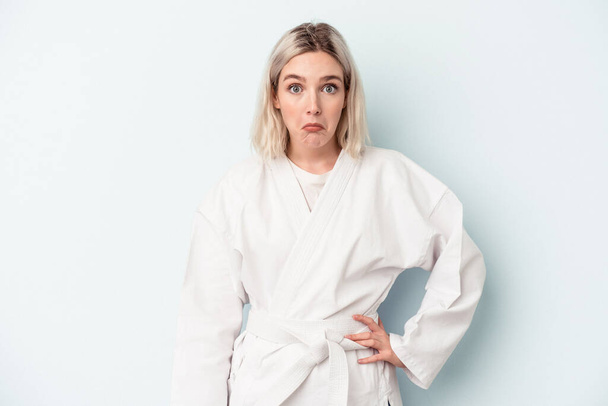 Young caucasian woman doing karate isolated on blue background shrugs shoulders and open eyes confused. - Photo, Image