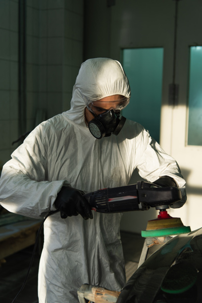 Workman in hazmat suit holding car polisher near part in garage  - Foto, imagen