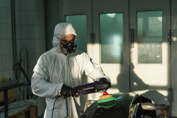 Workman in protective suit and respirator polishing car part in garage  - Photo, Image