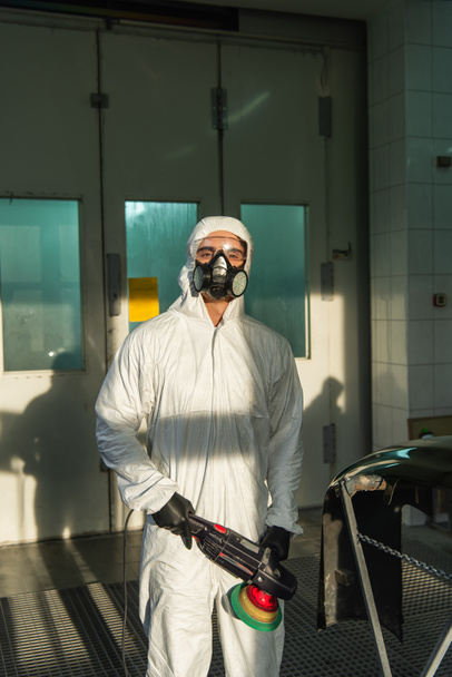 Workman in respirator and hazmat suit holding car polisher and looking at camera in garage  - Zdjęcie, obraz