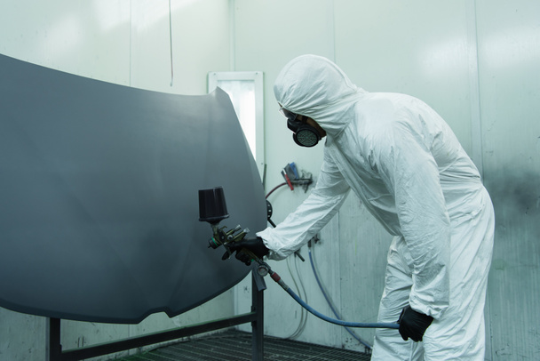 Workman in protective suit spraying varnish on car part in garage  - Foto, Bild