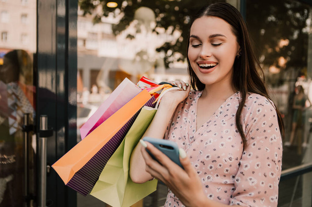 Shocked lady hold many bags and surfing internet with phone shopper woman dressed pink flower pattern dress carrying enjoying new clothes packs things after shopping buyings sales black friday concept - Foto, Imagen