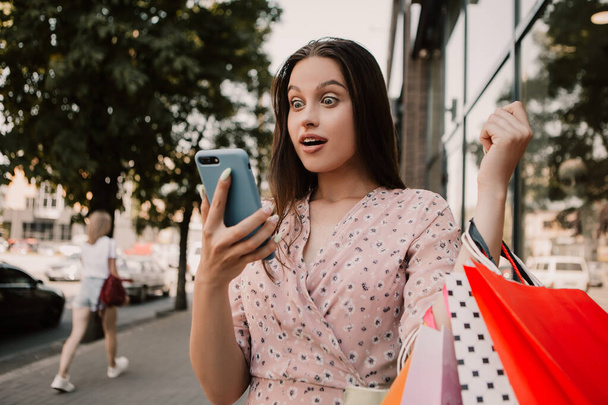 Shocked lady hold many bags and surfing internet with phone shopper woman dressed pink flower pattern dress carrying enjoying new clothes packs things after shopping buyings sales black friday concept - Фото, изображение