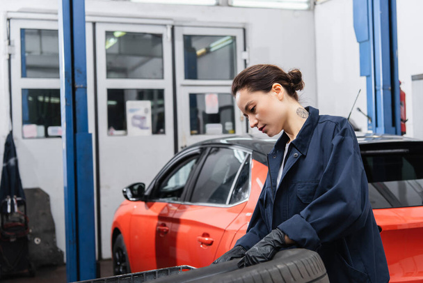 Femme de travail tatouée avec des gants tenant un pneu près d'une voiture floue dans un garage   - Photo, image