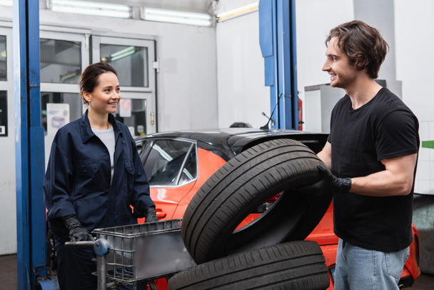 Mecánica sonriente de pie cerca de neumáticos y coche borroso en el garaje  - Foto, Imagen