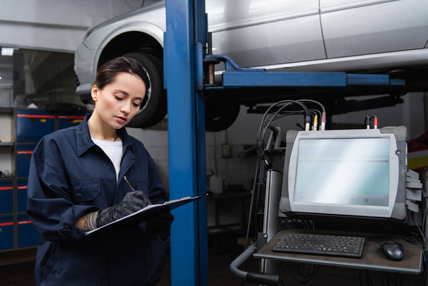 Workwoman writing on clipboard near computer and car in service  - Photo, Image