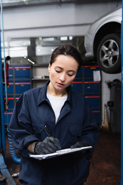 Trabajadora en guantes y escritura uniforme en portapapeles cerca de coche borroso en el garaje  - Foto, Imagen