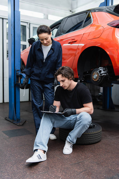 Young workman writing on clipboard on tire near colleague with screwdriver in car service  - Фото, изображение