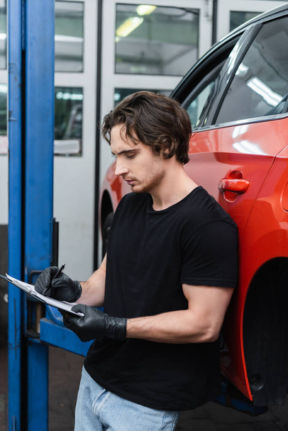 Young mechanic in gloves writing on clipboard near car in garage  - 写真・画像