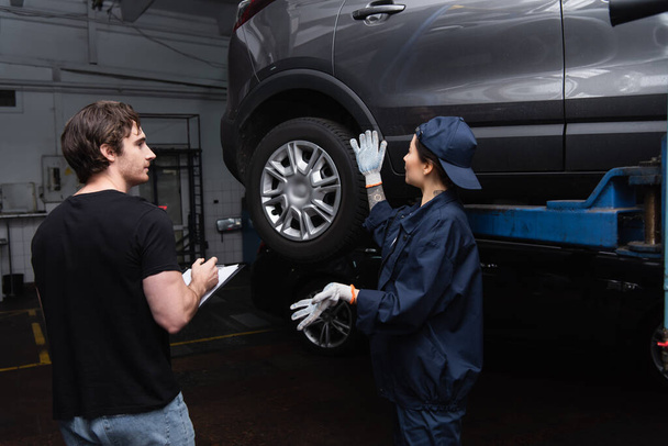 Mechanic pointing at car near colleague with clipboard in garage  - Foto, Imagem