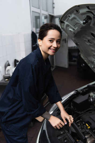 Smiling mechanic looking at camera near car in garage  - Фото, изображение