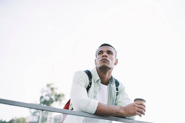 From below of thoughtful young black guy in casual clothes with backpack leaning on glass fence on city street with cup of drink in sunny summer day under cloudless sky - Fotó, kép
