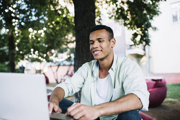Cheerful young black male freelancer sitting in comfort cafeteria located in park and working on modern laptop on summer sunny day - Valokuva, kuva