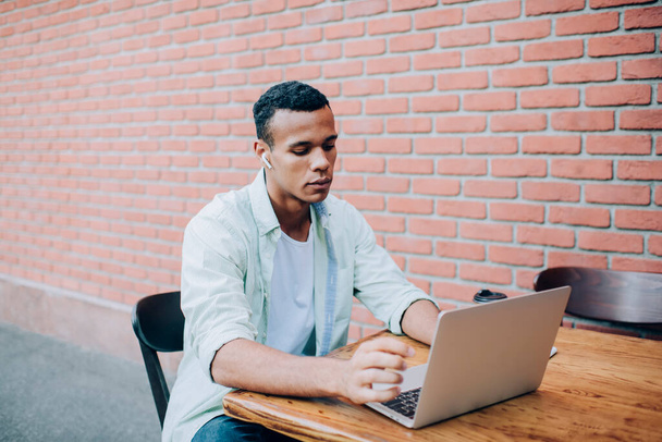 Confident young black man in casual outfit sitting on stool at table while working remotely on netbook with earphones in street in summer day near brick wall - Φωτογραφία, εικόνα