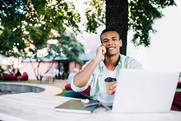 Young cheerful African American male freelancer with toothy smile looking at camera while sitting on street with coffee and laptop talking on smartphone - Fotografie, Obrázek