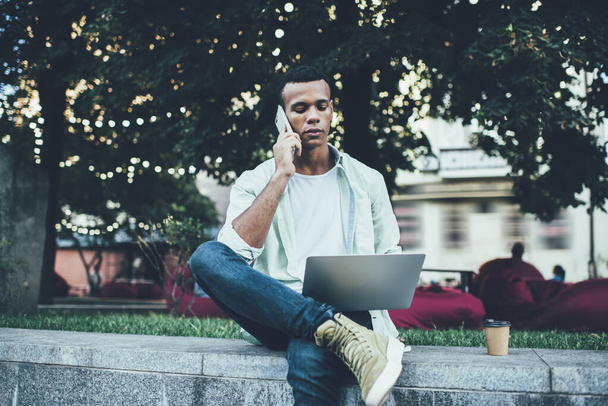 Young pensive African American male browsing netbook while discussing business on smartphone on concrete parapet near lawn with green grass - Fotó, kép