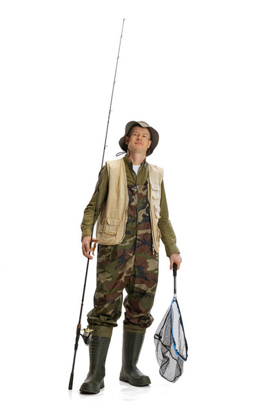 Portrait of young man, professional fisherman with fishing rod, spinning and equipment going to river isolated over white studio background - Photo, Image