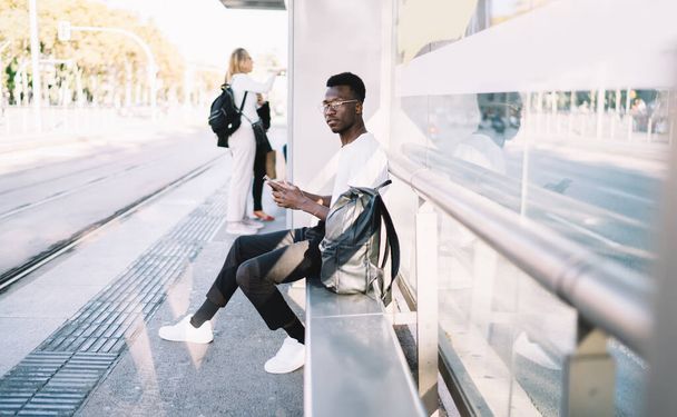 Side view of black man in casual outfit and eyeglasses sitting on metal bench with rucksack on tram station in sunlight while using cellphone and looking at camera - Φωτογραφία, εικόνα