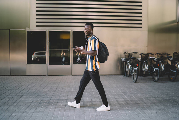 Full body side view young African American student in casual clothes with backpack and earphones walking along modern city district while using smartphone and smiling at camera - Foto, immagini