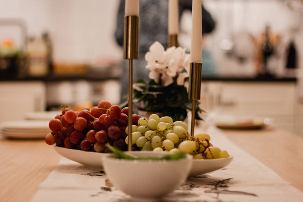 Εορταστικό τραπέζι με αναμμένα κεριά χρυσό κηροπήγιο. Κόκκινα και πράσινα σταφύλια σε λευκό μπολ. - Φωτογραφία, εικόνα