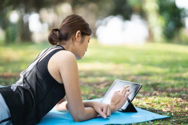 Asian fitness woman doing a yoga according to a video clip. Asian woman using tablet and doing yoga in park - 写真・画像
