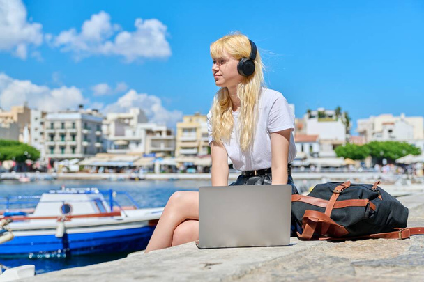 Adolescente donna in cuffia con computer portatile seduto vicino all'acqua, porto marittimo con barche - Foto, immagini