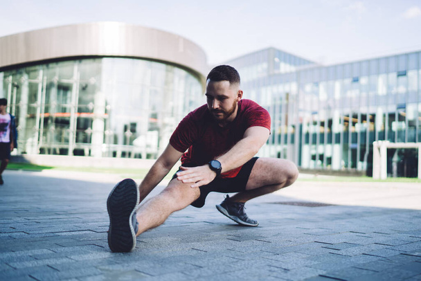 Sporty male in activewear looking down stretching legs while warming up before workout on street near modern glass building in cloudless weather - Фото, изображение
