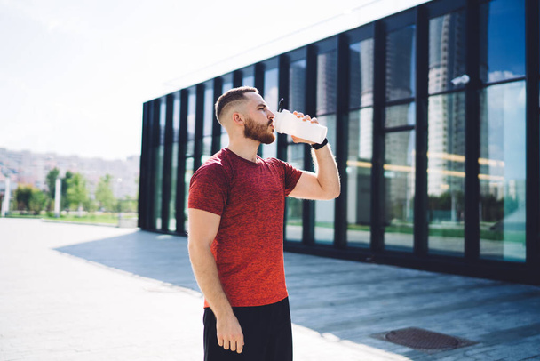 Side view of bearded male in sportswear drinking water from bottle after training while standing near building with glass walls - Foto, Imagem