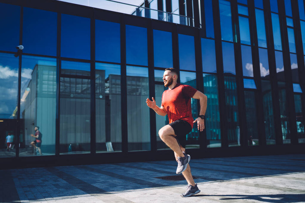 Full body of determined man in sneakers doing exercises on pavement near glass office building while living healthy lifestyle and looking away - 写真・画像