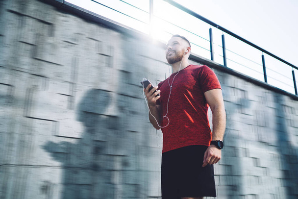 From below of smiling male in red t shirt and shorts looking away using smartphone and earphones on street near stone wall - Fotoğraf, Görsel