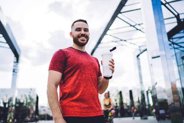 From below of smiling bearded male athlete with bottle of water looking at camera and chilling after training while spending time in city center - Foto, afbeelding