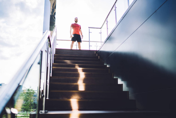 From below of young male in activewear standing on stairs of contemporary building while preparing for workout in modern city - Foto, imagen