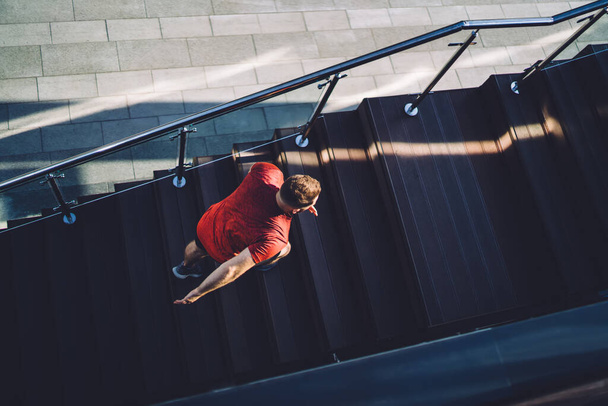From above of full body anonymous male athlete in red tee shirt doing exercises in city while running upstairs during sunny morning - 写真・画像