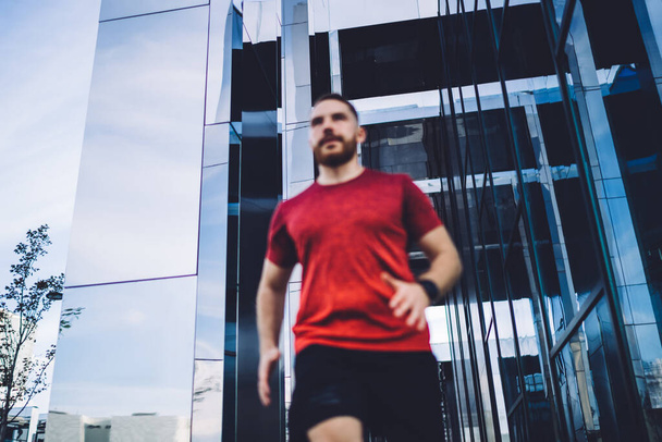Low angle of determined male in sportswear jogging in city near contemporary building during cardio exercise in sunny summer day - Fotoğraf, Görsel