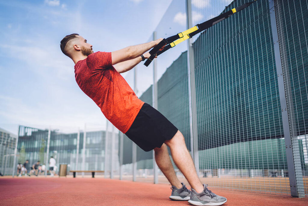 Side view of full length male athlete in sportswear doing exercises with suspension straps while standing near fence on sports ground on summer day - Fotó, kép