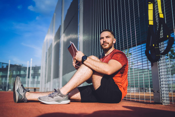 Low angle of focused guy sitting near net fence and listening to lecture in earphones while preparing for sprint outside - Foto, Bild